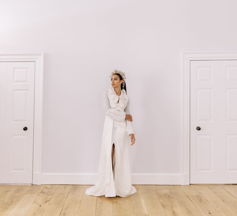 Bride stands in bright white room whilst wearing bridal gown with ruffled front and leg split