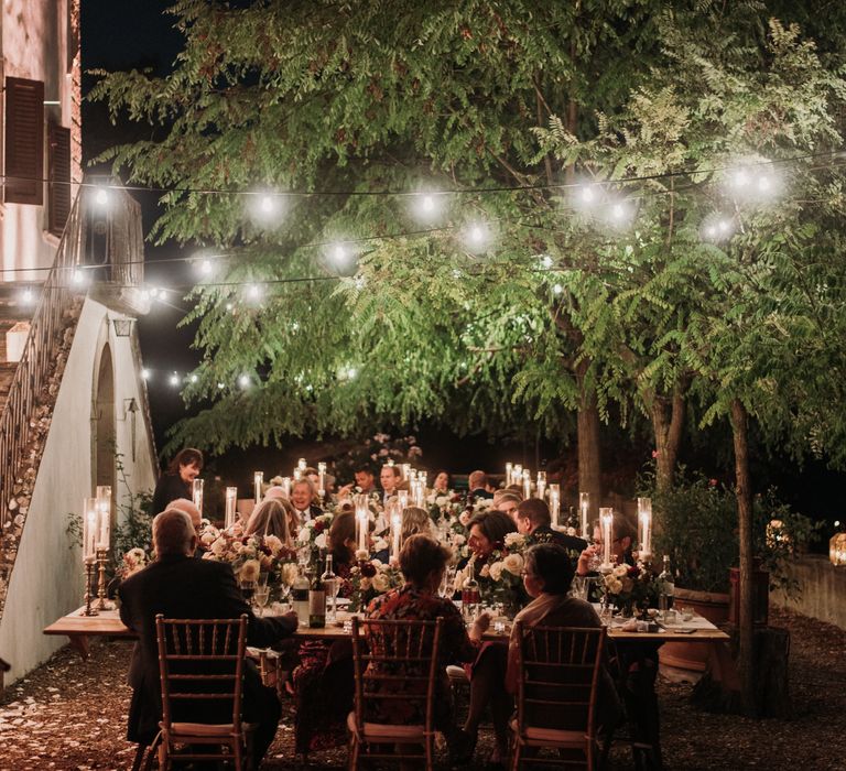 Guests sit to dinner during outdoor wedding reception with candlelights and twinkling fairy lights above