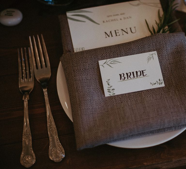 Bride table place name with botanical details on grey napkin with rosemary sprig, silver cutlery and diamond cut glass tumbler