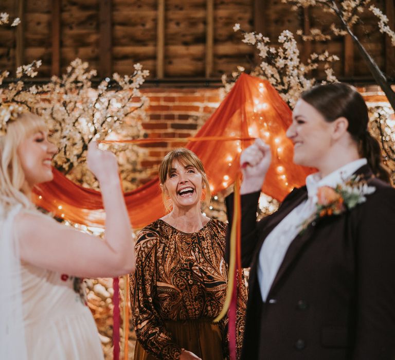 Two brides in front of blossom trees draped with orange fabric, holding up orange ribbons in front of a celebrant for pumpkin spice wedding