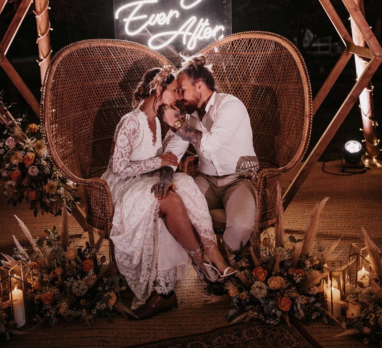 Boho bride and groom sitting on wicker peacock chairs under a neon Happily Ever After wedding sign 