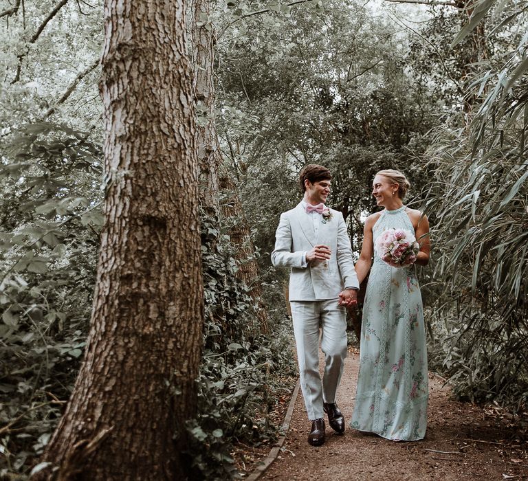 Bride & groom walk together through woodland whilst holding hands on the day of their wedding