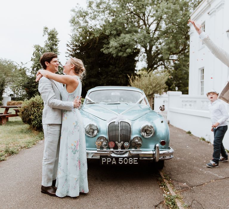 Bride & groom stand beside pale blue vintage car on the day of their wedding as guest throws confetti around them