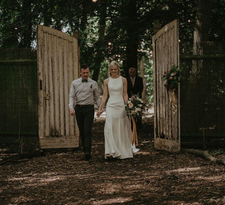 Bride & groom walk through large wooden doors on woodland pathway as bride holds floral bouquet