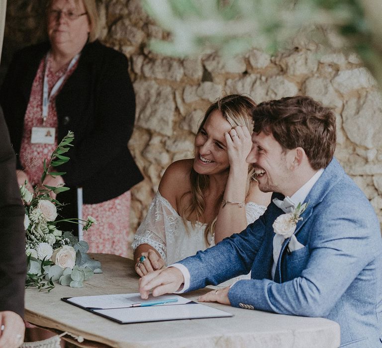 Bride in Grace Loves Lace Bardot wedding dress sits signing marriage registry with groom in blue suit with floral buttonhole at Isle of Wight wedding with macrame wedding decor