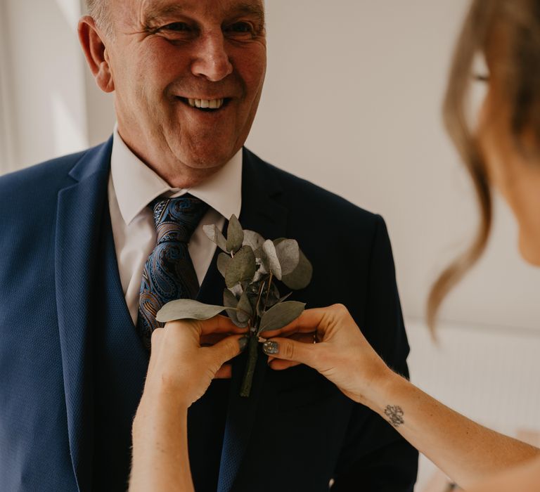 Bride applies floral buttonhole on father's blazer on her wedding day as he smiles | Mark Bamforth Photography