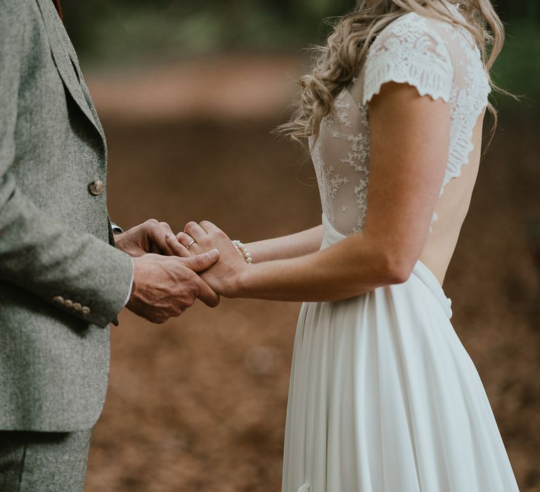 Bride in white lace cap sleeve open back wedding dress holds hands with groom in grey herringbone suit during woodland wedding ceremony in Norfolk