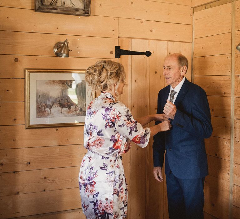 Bride in floral sating dressing gown with curled wedding ups adjusts suit of father of the bride in wooden room before tip wedding at Inkersall Grange Farm