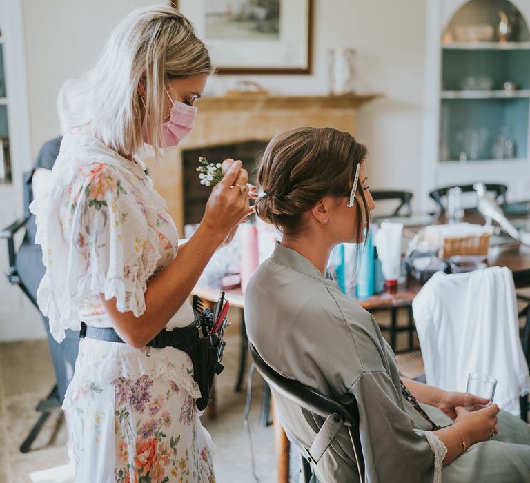 Bride in mint green satin lace trimmed dressing gown sits in chair as she has her hair done by woman in white floral dress before summer wedding at Primrose Hill Farm