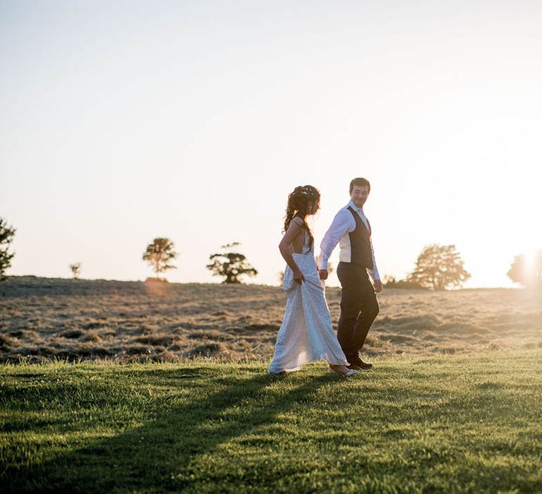 Bride & groom walk across green grass as the sun begins to set