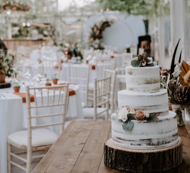 White three tier semi-naked wedding cake on tree slice cake stand with foliage decor on wooden table for marquee wedding reception in Bedfordshie