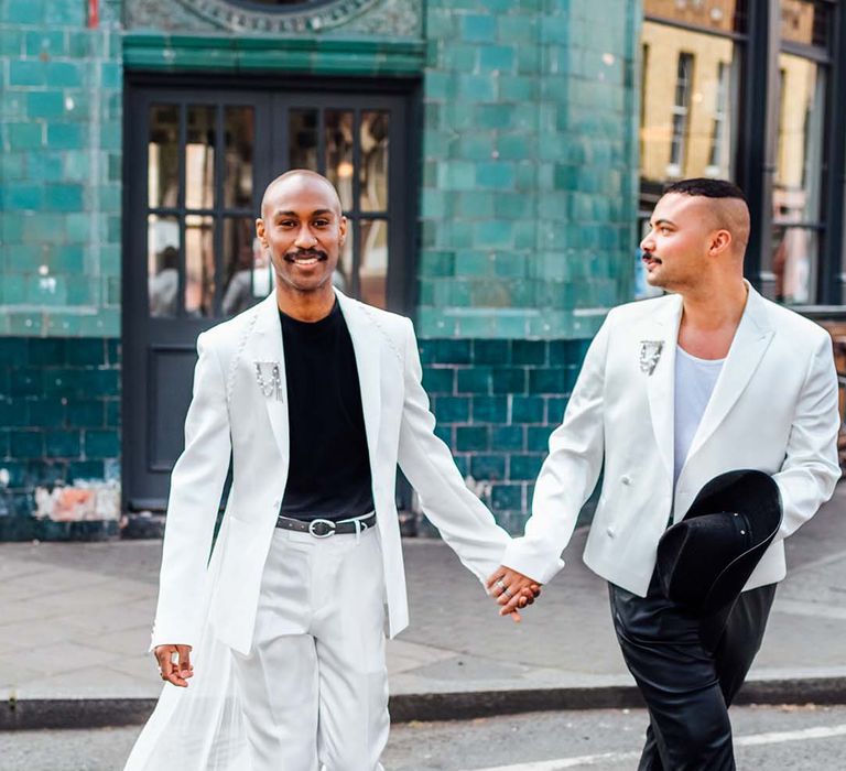 two stylish grooms in a white satin suit, leather trousers and double breasted jacket holding hands 