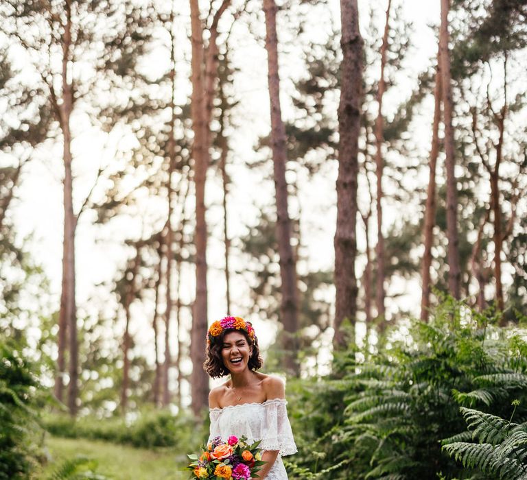 Bride in off the shoulder Grace Loves Lace wedding dress and flower crown holding colourful bridal bouquet stands in woodland after outdoor wedding ceremony at The Bridal Barn