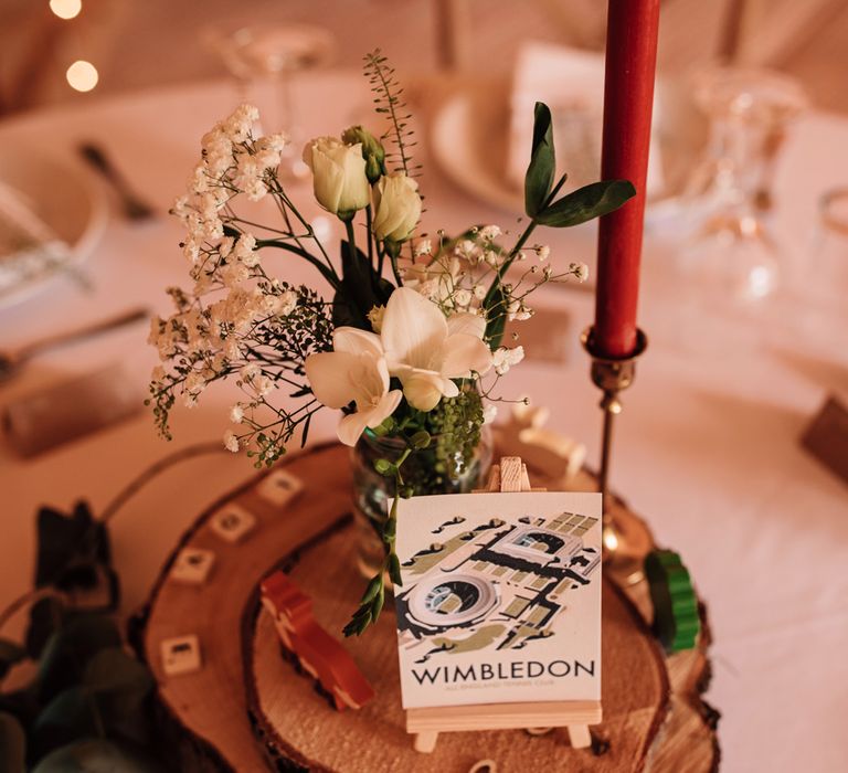 Rustic wedding centrepiece with burgundy candle, glass jar of white and green florals and Wimbledon postcard