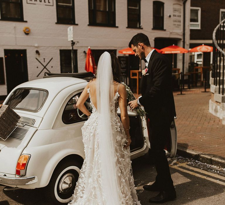 Bride wears Alena Leena Bridal gown complete with appliqué design and carries floral bouquet whilst entering vintage Fiat 500