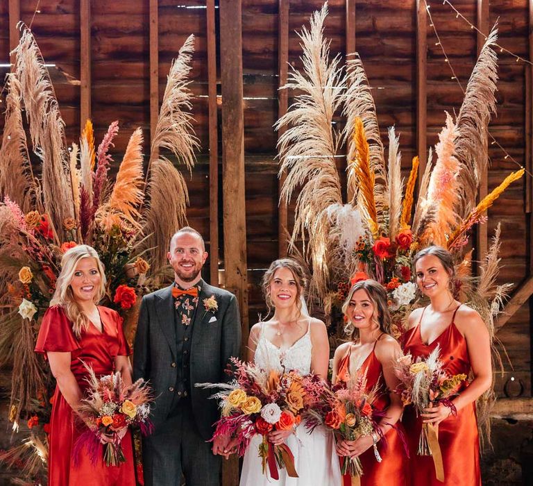 Bride in lace wedding dress holding dried flower bouquet stands with groom in green suit and patterned shirt and bridesmaids in orange satin bridesmaid dresses in front of pampas grass floral installation 