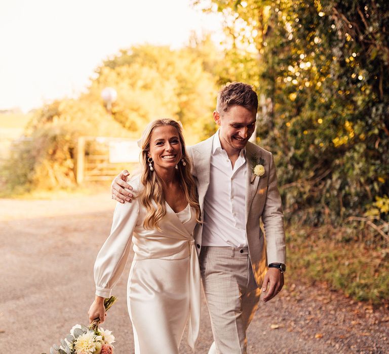 Bride in white The Own Studio wedding dress with satin wrap top and silver heeled sandals walks holding colourful bouquet with groom in open white shirt and light grey suit 