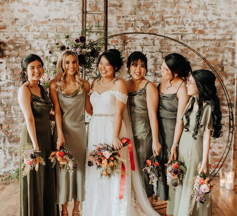 Bride in bardot wedding dress stands with bridesmaids in sage green satin bridesmaid dresses in front of floral moongate all holding mixed wedding bouquets with foliage and florals at industrial wedding venue in Liverpool