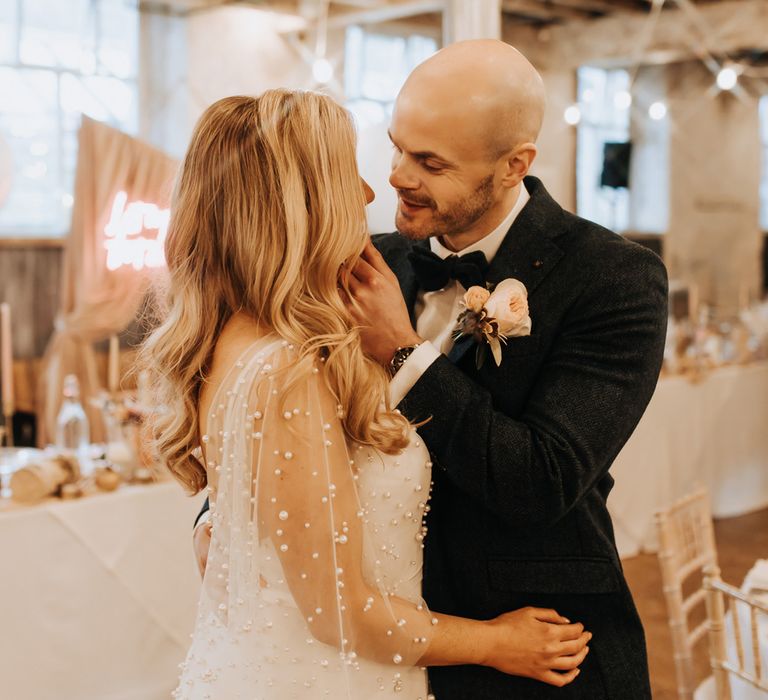 Groom in navy tweed suit with floral buttonhole stands with bride in mesh sleeve wedding dress with pearls at wedding reception