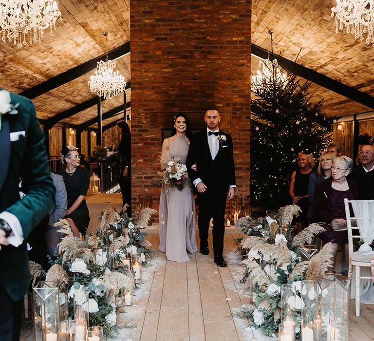 Bridesmaid in taupe dress and groomsman in black tie walk down the aisle together with chandeliers and Christmas tree decor for winter wedding 