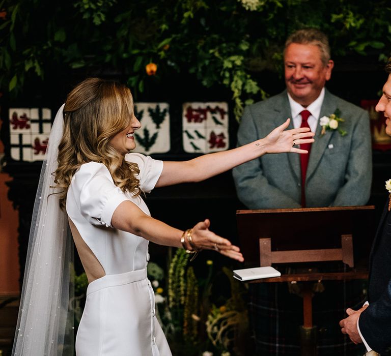 Bride with pearl veil opens her arms wide for the groom at the altar 