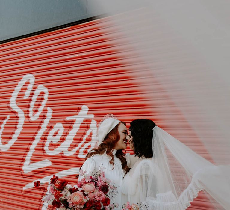 Two brides kissing in a short wedding dress long veil wedding outfit and berret hat, high neck wedding dress with long sleeves holding a red and pink wedding bouquet