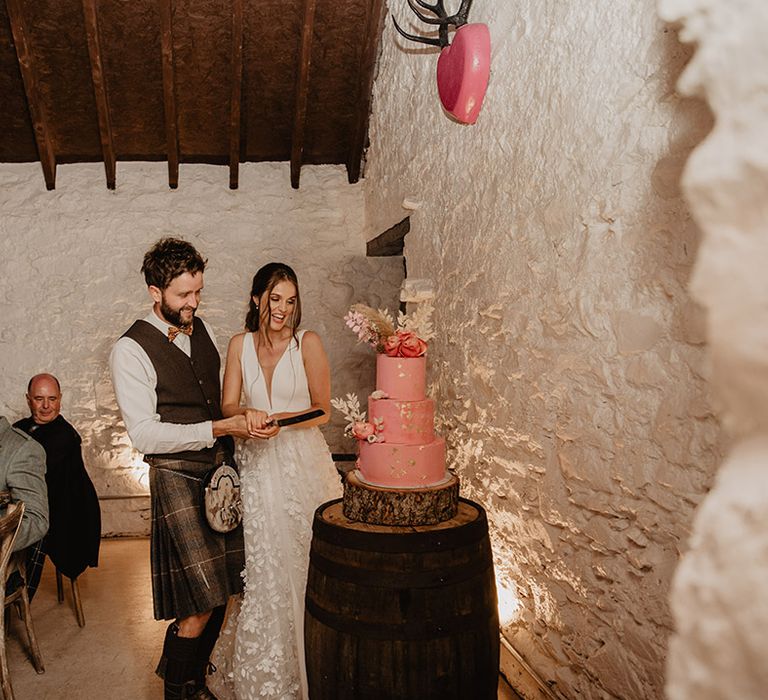 Bride and groom cut into their pink three tier wedding cake with gold flakes and pink and white dried flower decorations