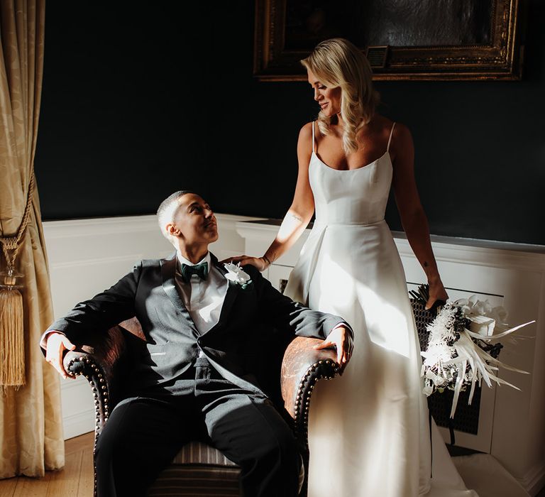 Blonde bride in a princess wedding dress with spahetti straps holding an orchid wedding boqueut next to her partner sitting in a tuxedo and bow tie 