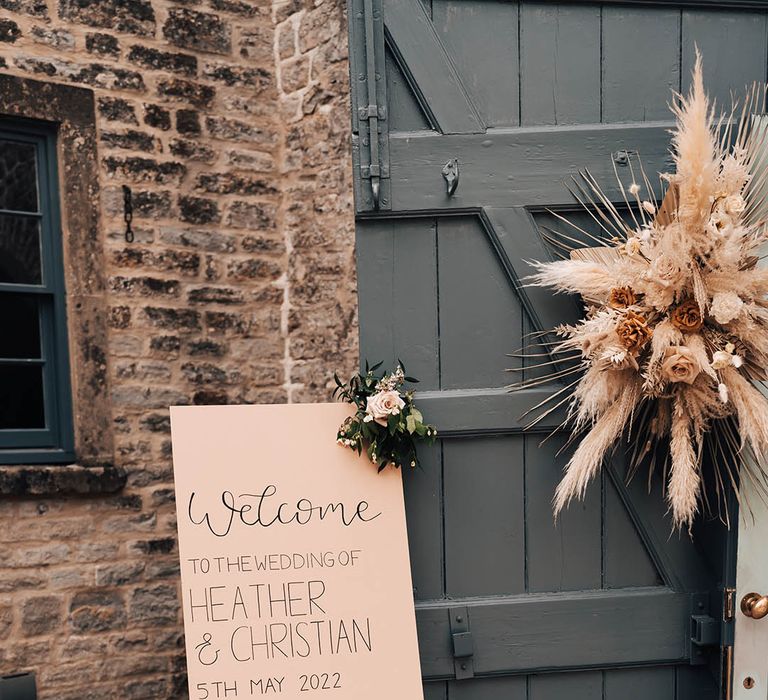 Beige and black welcome wedding sign with small order of the day sign next to large dried flower display and candles in glass lanterns