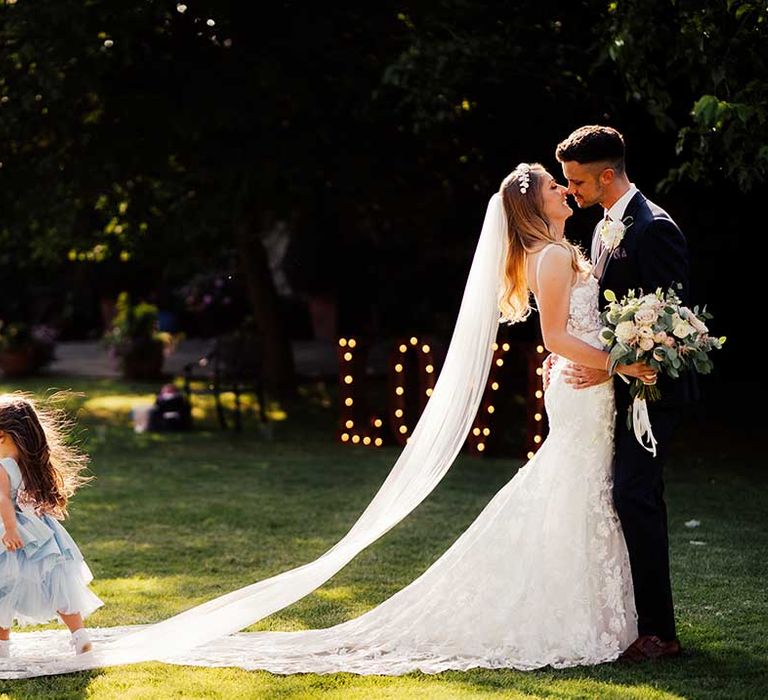 Bride and groom lean in for a kiss together with young wedding guest in blue dress runs over the bride's train and veil