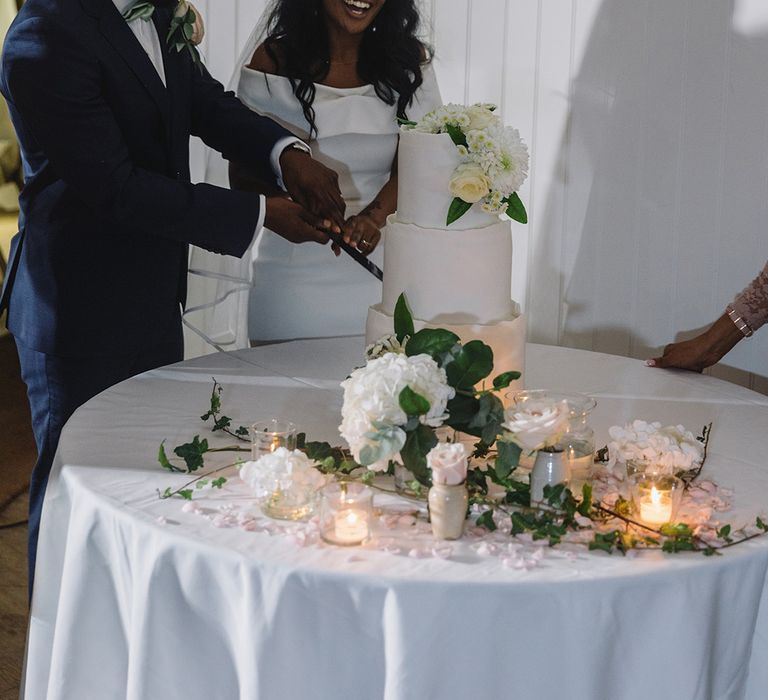 Bride and groom cut into their three tier white wedding cake decorated with white flowers