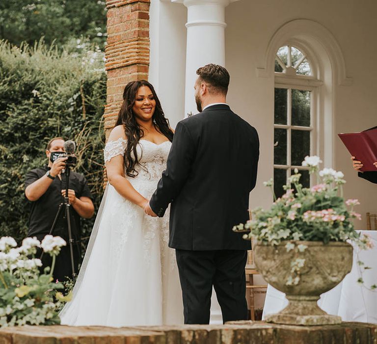 Bride and groom hold hands and smile at each other as the celebrant reads out to them 