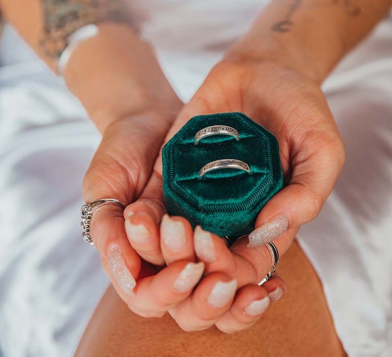 Bride holds engraved wedding bands in green velvet ring box with glittery wedding nails