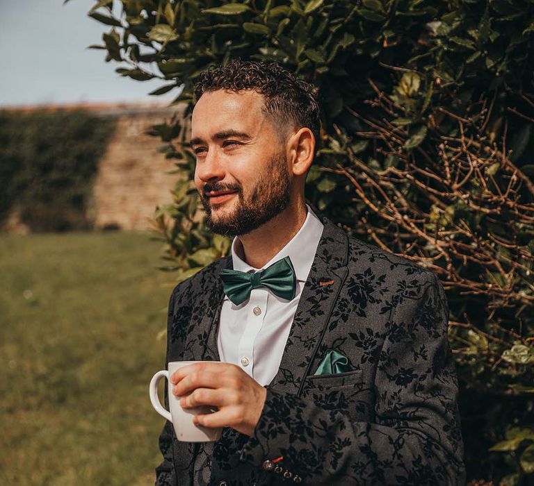 Groom in three piece black patterned suit with dark green bow tie holding onto white mug 