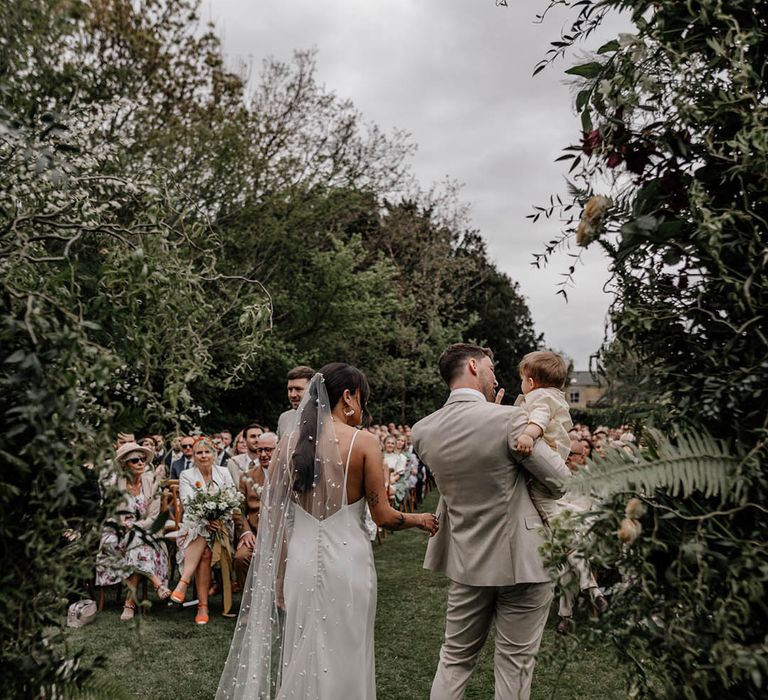 Bride in a satin wedding dress and bespoke pearl veil and groom in a beige suit holding their son at their outdoor garden wedding ceremony