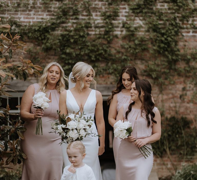 Bride in deep v wedding dress with pearl hair pins walks with bridesmaids in pink dresses and daughter in white pearl cardigan and tulle dress 