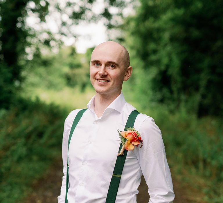 Groom poses outdoors wearing white shirt, green braces and brightly coloured floral buttonhole 