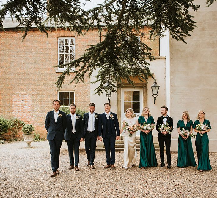 Bride & groom stand with bridesmaids who wear emerald green satin bridesmaid dresses and groomsmen in blue blazers and white shirts