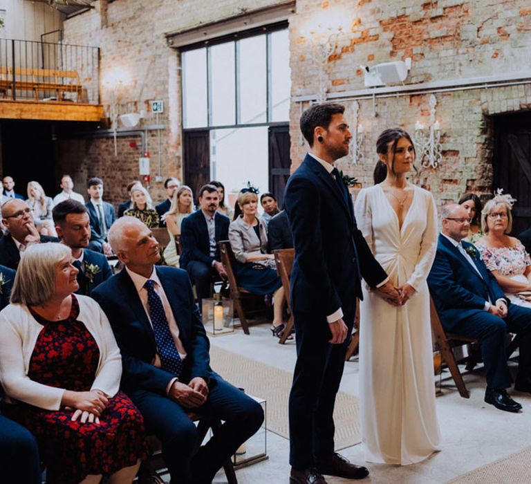 Bride and groom stand holding hands for their ceremony with wedding guests watching on 