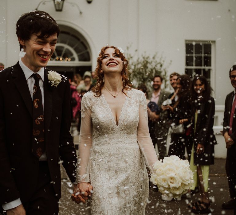 Groom wearing brown suit with psychedelic tie leaves Belair House with his bride in lace wedding dress carrying white rose bouquet as confetti falls around them 