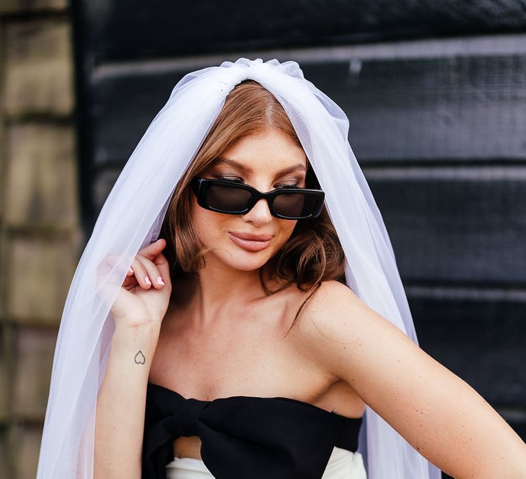 Bride wears sunglasses and cathedral veil with white mini dress complete with large black bow to front  