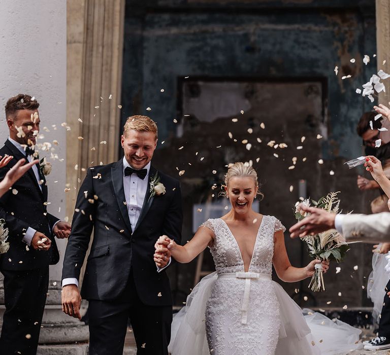 Groom walks down staircase with his bride wearing Eva Lendal wedding dress with detachable skirt as wedding guests throw confetti around them