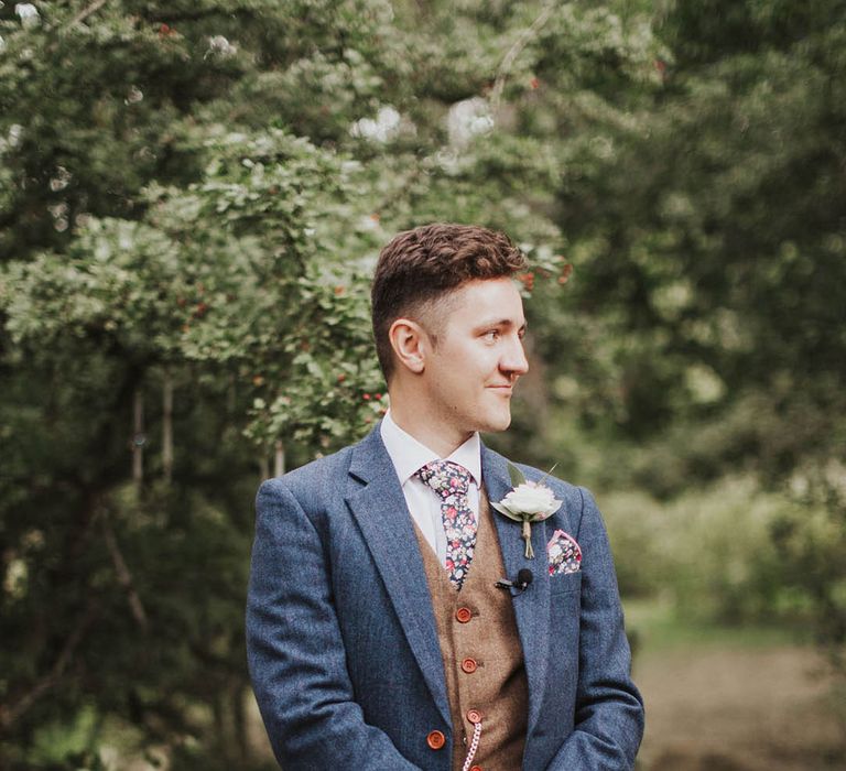 Groom stands and gets the first look at the bride holding his vows as he wears in a blue suit with a floral tie and handkerchief with a brown waistcoat 