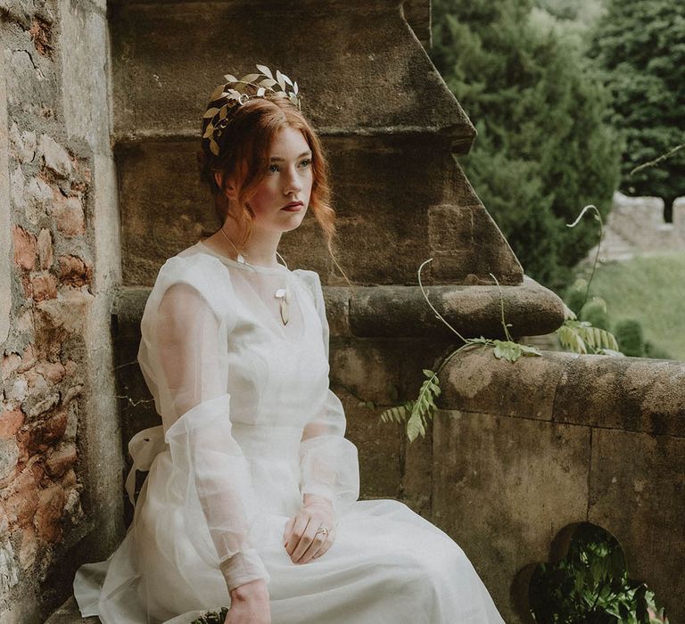 Bride with red hair sitting on a step at Bishop's Palace Wells in a two layer wedding dress and gold leaf crown holding a handed wedding bouquet 