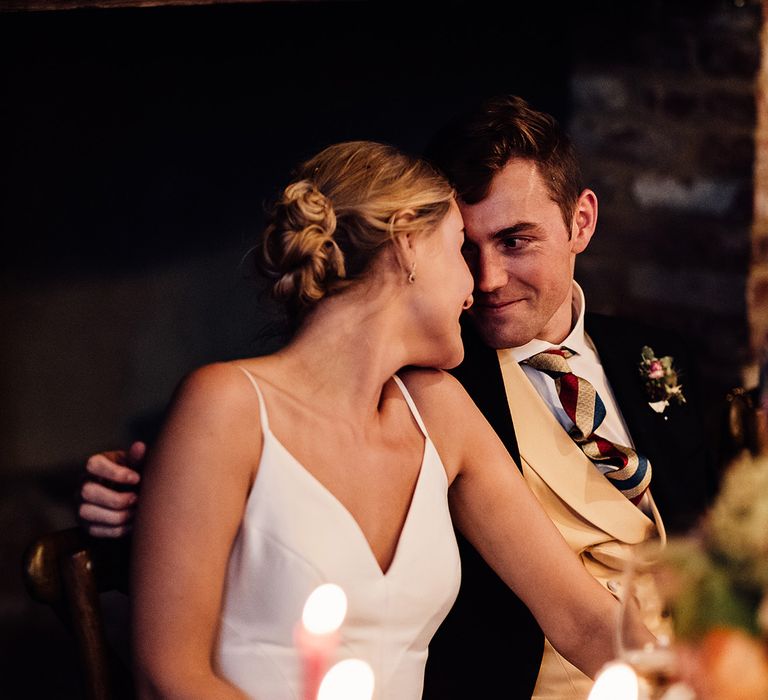 Bride and groom sit for their wedding reception and reach over to touch each other and smile 