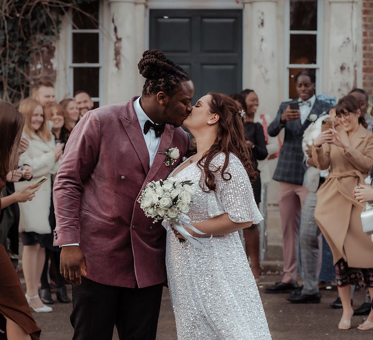 Bride in a short sleeve sequinned wedding dress kisses her groom in a pink jacket 