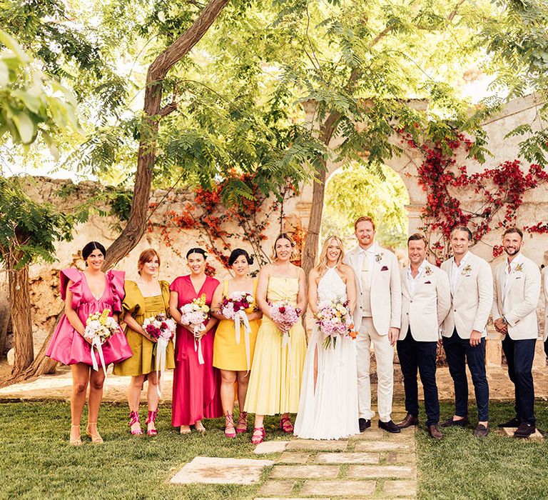 Bride and groom stands with their wedding party in bright and colourful bridesmaid dresses and cream groomsmen suits