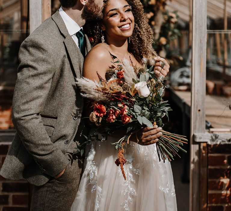 Groom in a grey tweed suit kisses the bride on the forehead who carries an autumnal bouquet with pampas grass 