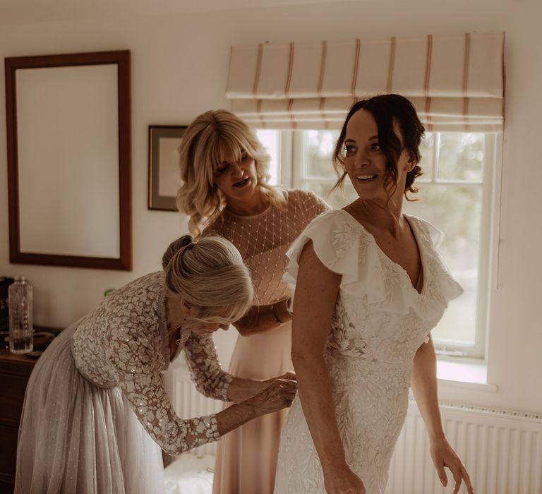 Bridesmaids in a blush pink pearl dress and mother of the bride helps the bride into her bespoke Emma Beaumont Atelier wedding dress 