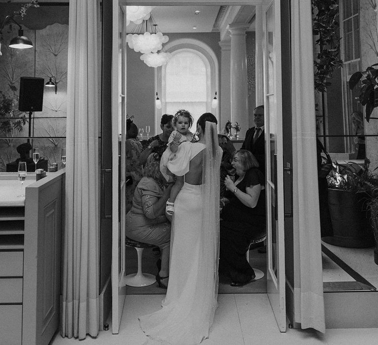 Bride in open back puff sleeve wedding dress with long pearl detailed veil holding baby at Somerset House 